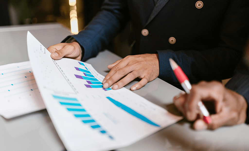Mãos analisando documentos com gráficos de barras coloridos sobre uma mesa, simbolizando a avaliação de dados e performance empresarial.