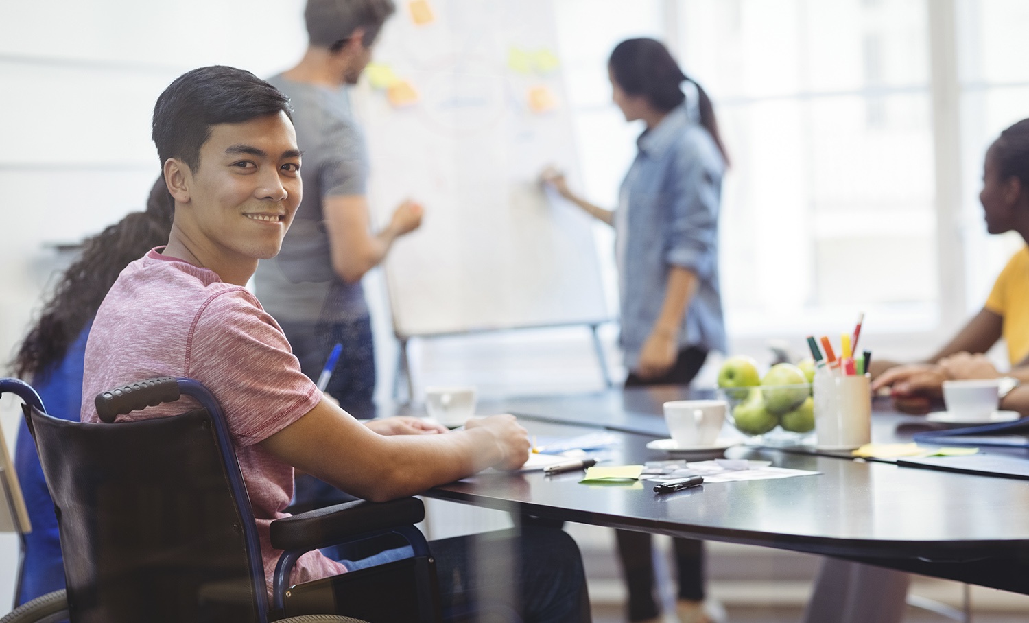 Homem em cadeira de rodas sorrindo em reunião corporativa, com colegas diversos ao fundo. Ilustra a diversidade no ambiente de trabalho.