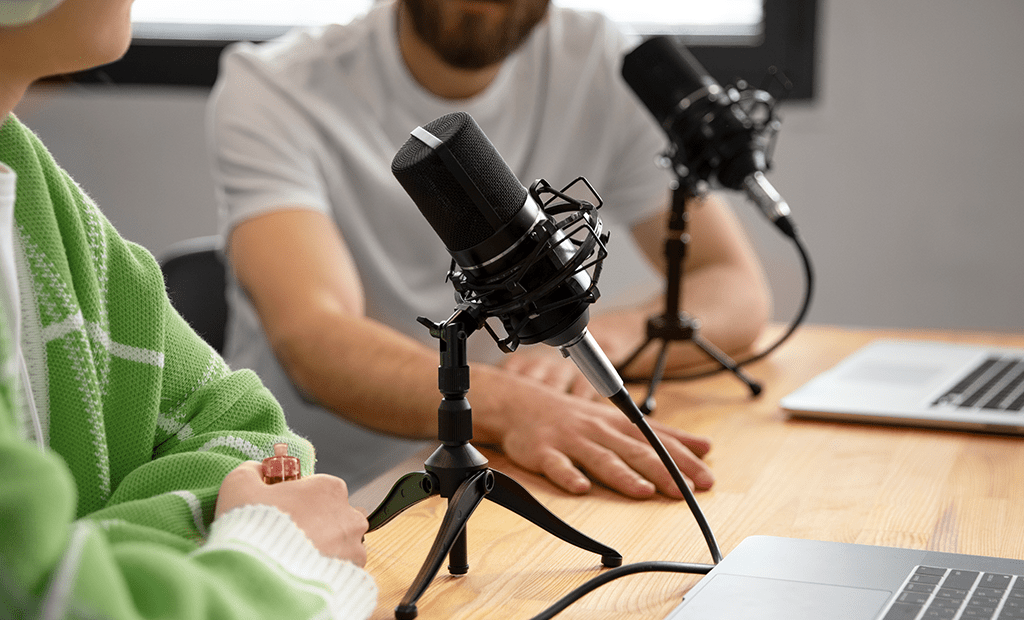 Dois pessoas gravam podcast com microfones profissionais e laptops sobre uma mesa de madeira, ilustrando o conteúdo do episódio da EqualWeb.