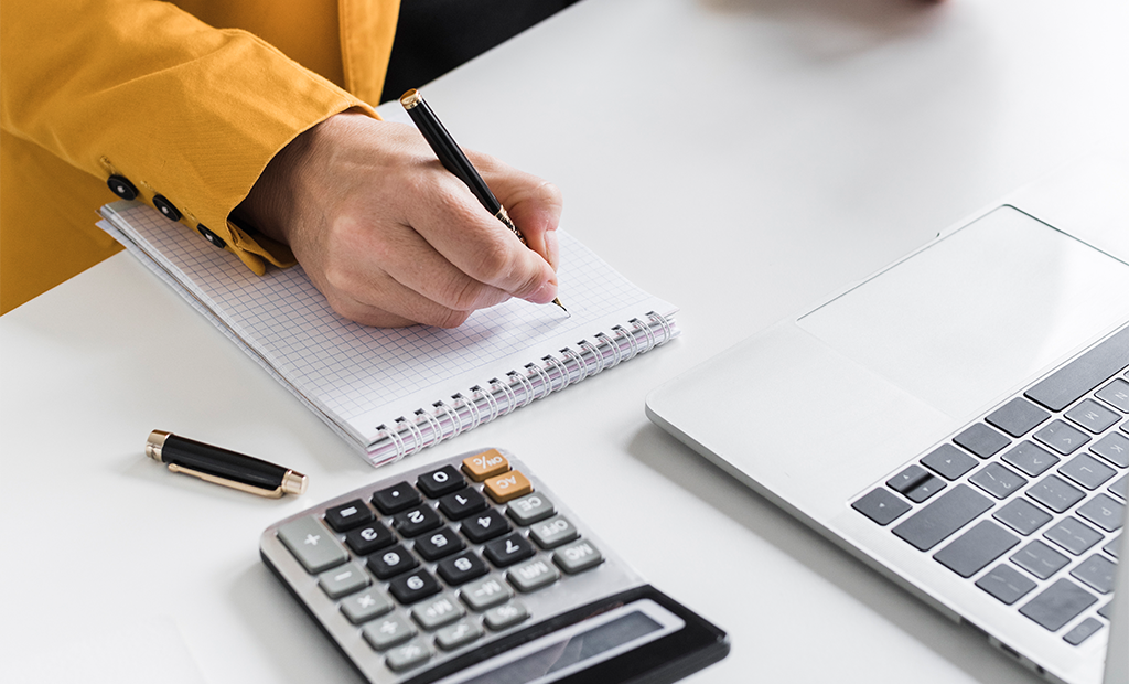 ROI da acessibilidade. Pessoa escrevendo em caderno quadriculado ao lado de notebook e calculadora sobre mesa de trabalho.