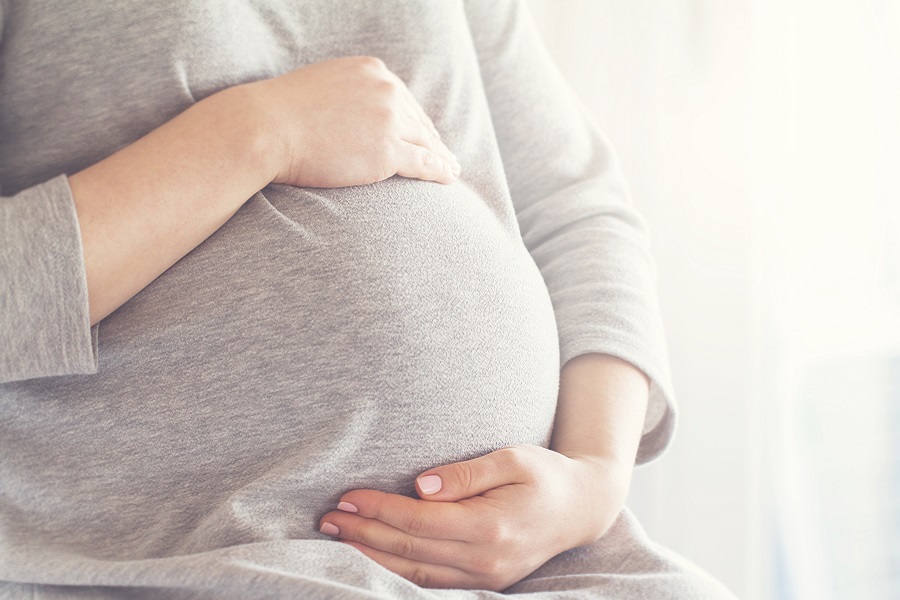 Tronco de mulher grávida em blusa cinza, com as mãos apoiadas na barriga. Ilustra o tema da gestação e testes de gravidez.