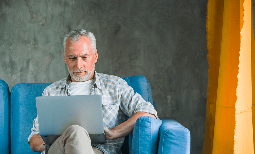acessibilidade digital. Imagem de um homem com cabelos e barbas grisalhas, sentado em um sofá azul mexendo em um notebook.