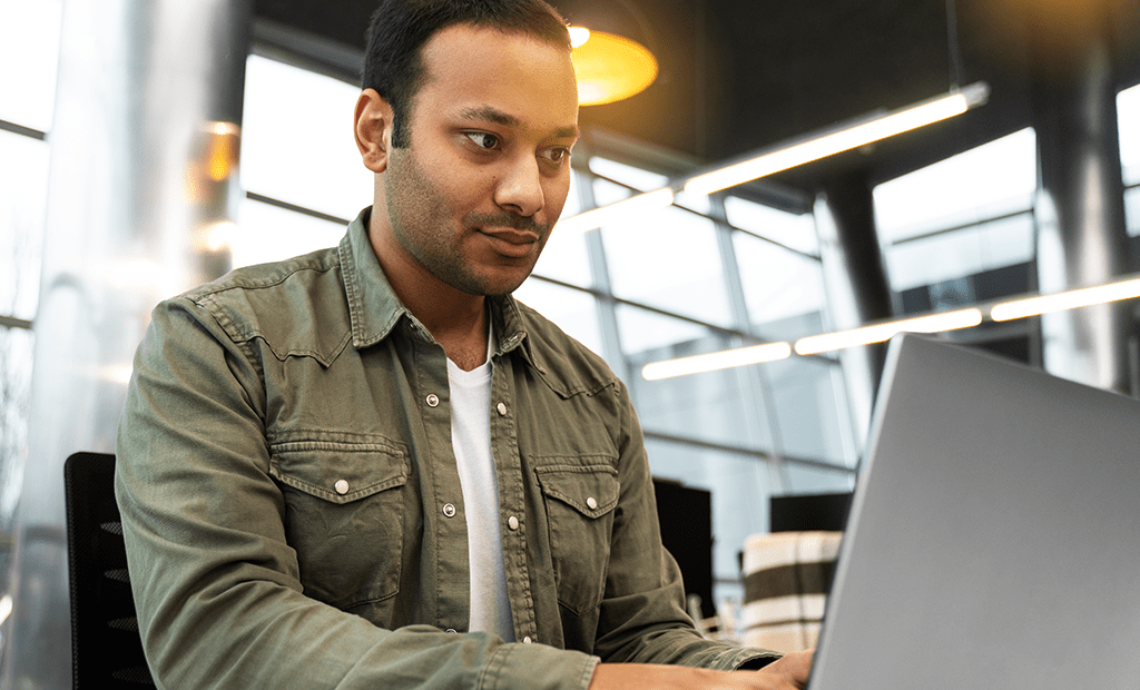  WCAG. Imagem de um homem negro, usando um casaco verde. Ele esta sentado em uma sala de reunião mexendo no computador.