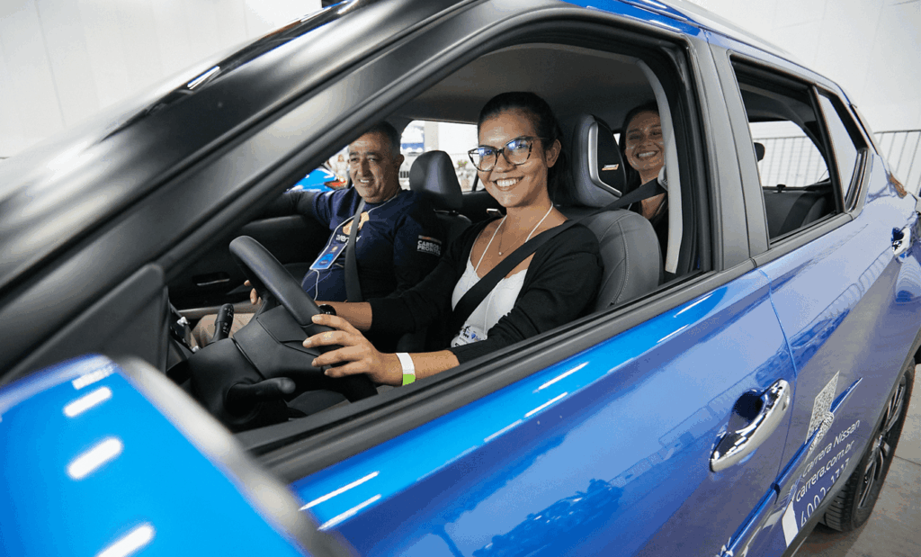 Reatech. Uma mulher morena de cabelo preso, óculos e casaco preto dentro de um carro adpatado, no banco do carona um homem branco de blusa azul e no banco traseiro uma mulher sorrindo.