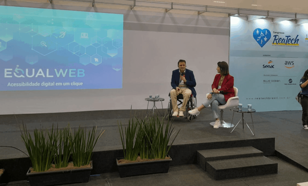 Reatech. No palco temos o Fabio Uzunof, homem branco, de cabelos castanhos e barba grisalha, vestindo calça bege, camisa branca e blazer azul, sentado em sua cadeira de rodas com um microfone na mão. Ao lado dele, temos a Diretora de Operações Marketplace na Via Varejo, Fernanda Winck, mulher branca, de cabelos castanhos em tamanho curto, vestindo calça jeans, camisa bege e casaco vermelho, sentada em uma cadeira branca. Ambos estão sorridentes olhando para a foto.