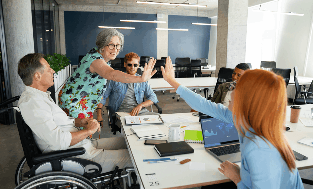 inlusão. A imagem é uma sala de reunião com alguma pessoas em volta da mesa, nela podemos observar uma menina ruiva próximo ao notebook tocando a mão de uma senhora de cabelos grisalhos que está usando um óculos ao lado temos também um cadeirante, um rapaz deficiente visual segurando uma bengala e um rapaz negro. 