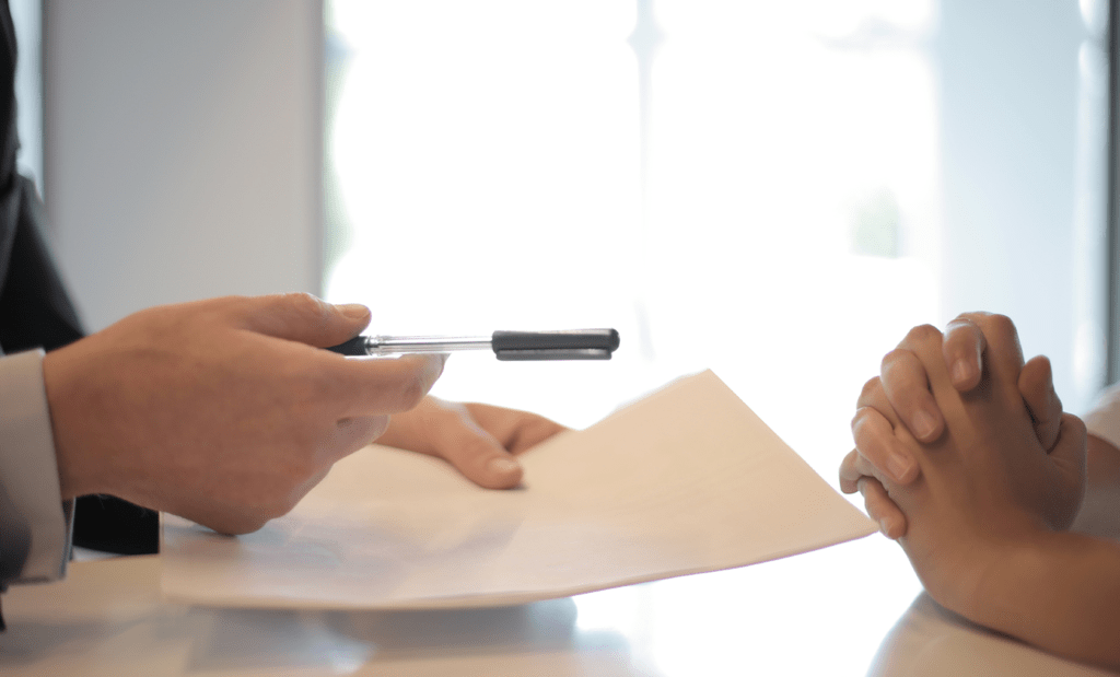 Equipe. Na imagem aparecem duas mãos sobre uma mesa, uma delas estão juntas com os dedos entrelaçados e a outra com uma das mão segura um documento e a outra uma caneta.