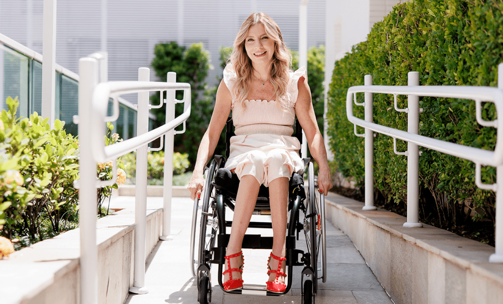 PCD. Imagem da Andrea Schwarz sentada na cadeira de rodas no meio de uma rampa de acesso, com um vestido branco e sapatos vermelhos, sorrindo. 
