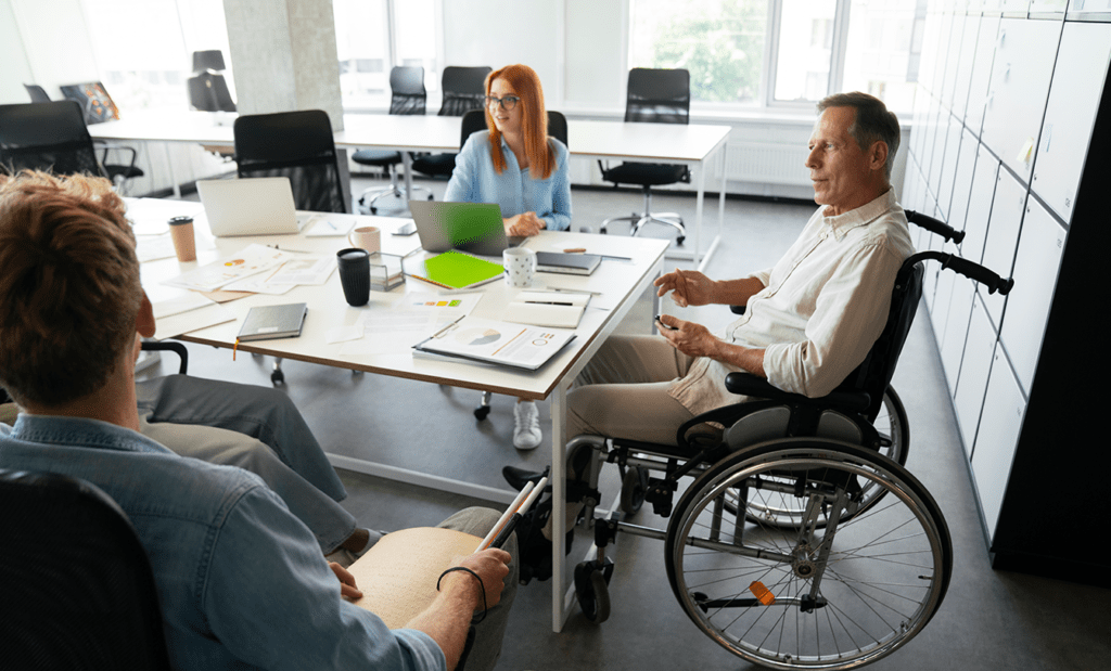 deficiência. Imagem de uma sala de reunião com três pessoas, uma jovem ruiva de óculos e roupa social, um homem cadeirante de roupa social e cabelos grisalhos e um jovem de roupa social. 