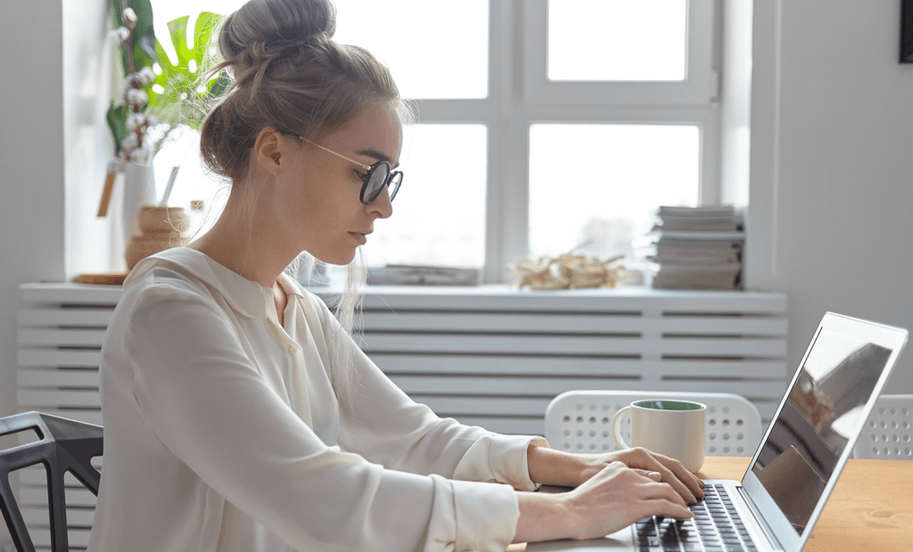 acessível. Uma menina branca, loira, usando um óculos de grau e mexendo no notebook ao lado está uma xícara de café. 