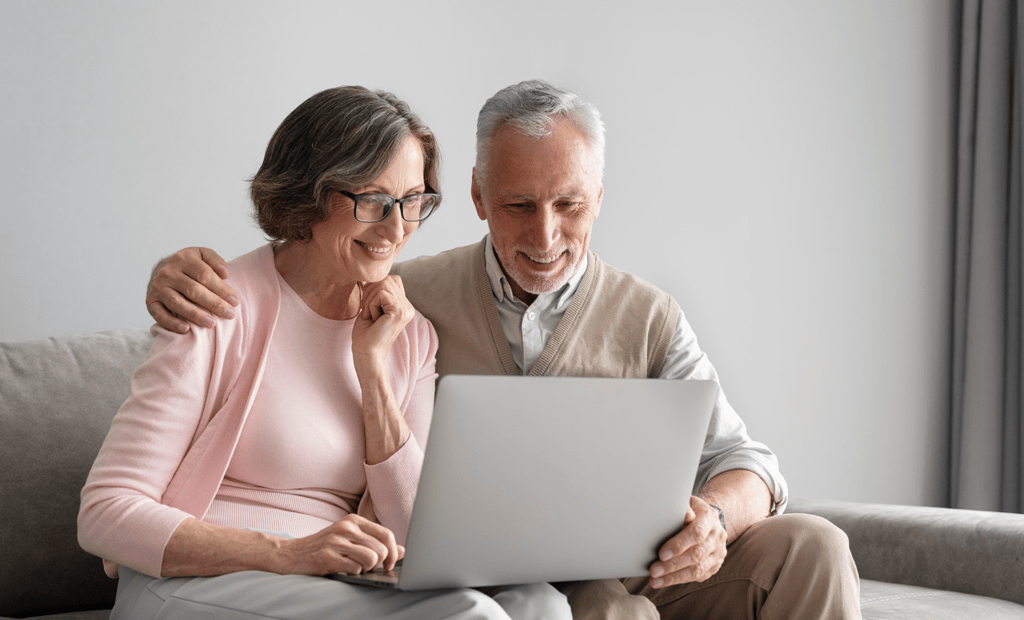 saúde. Um senhor e uma senhora de idade brancos e com cabelos grisalhos, sentados em frente ao notebook sorrindo para a tela.
