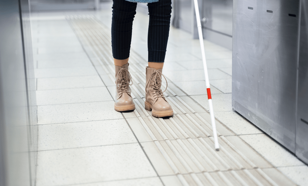 Conscientização. Imagem de uma pessoa andando sobre a sinalização para deficientes visuais no chão. Na imagem só aparece as pernas da pessoa e uma parte da bengala. 