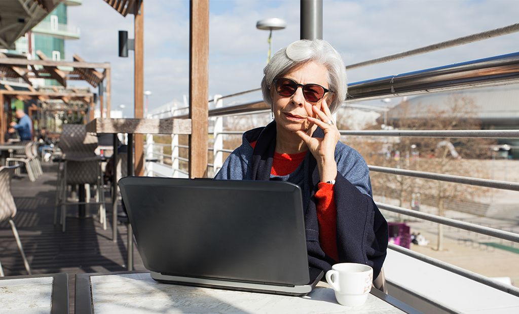 Turismo. Imagem de uma senhora de idade de cabelos brancos, sentada em uma mesa tomando um café e mexendo notebook.