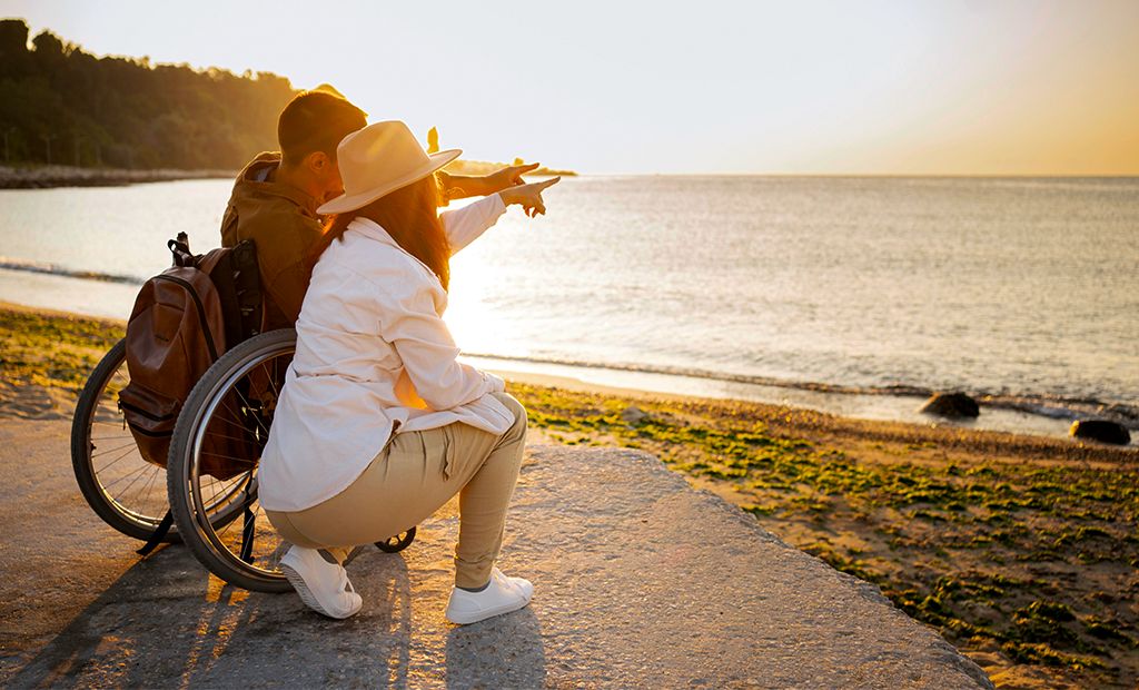 Turismo. Imagem de um casal na praia apontando para o horizonte. O homem está sentado na sua cadeira de rodas e a mulher está agachada ao lado dele.