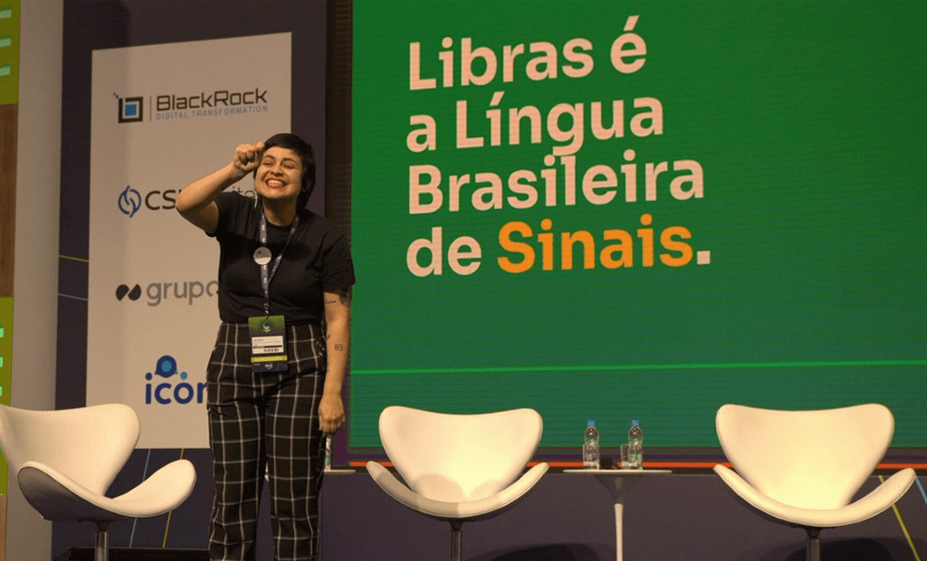 Transformação. Imagem de uma palestrante branca e de cabelos pretos e curtos em cima de um palco e no fundo aparece a seeguinte frase na tela: Libras é a Língua Brasileira de Sinais.