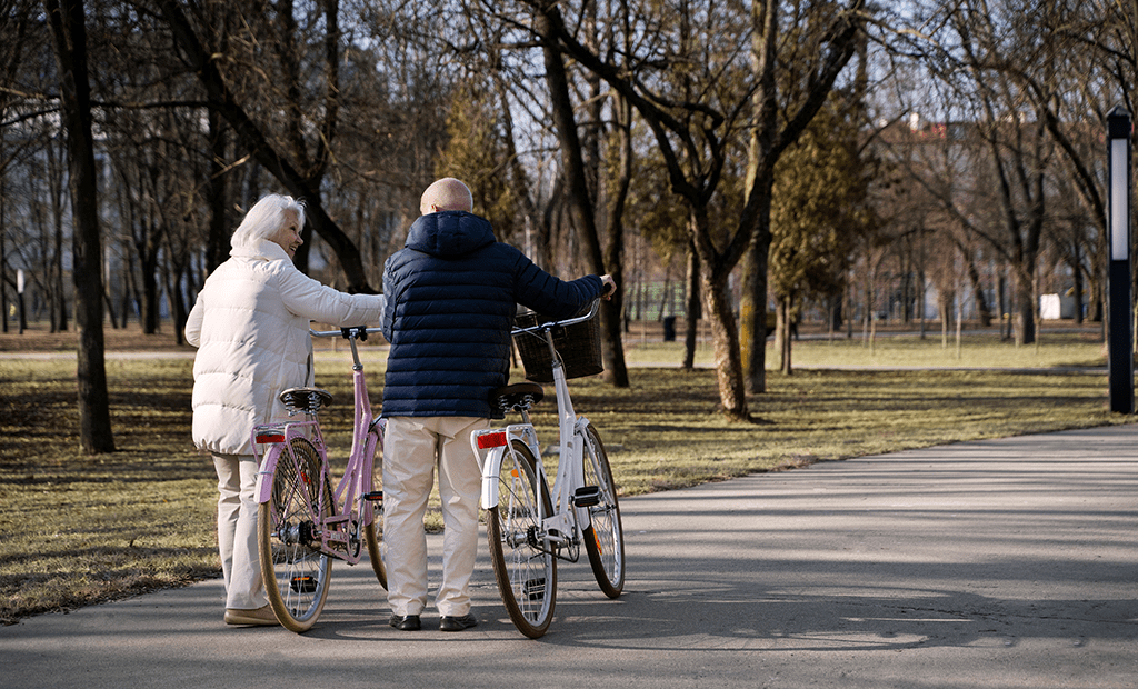 Acessibilidade. Imagem de um casal de idosos caminhando em um parque e empurrando uma bicicleta. O casal usa roupa de frio e caminha entre árvores.