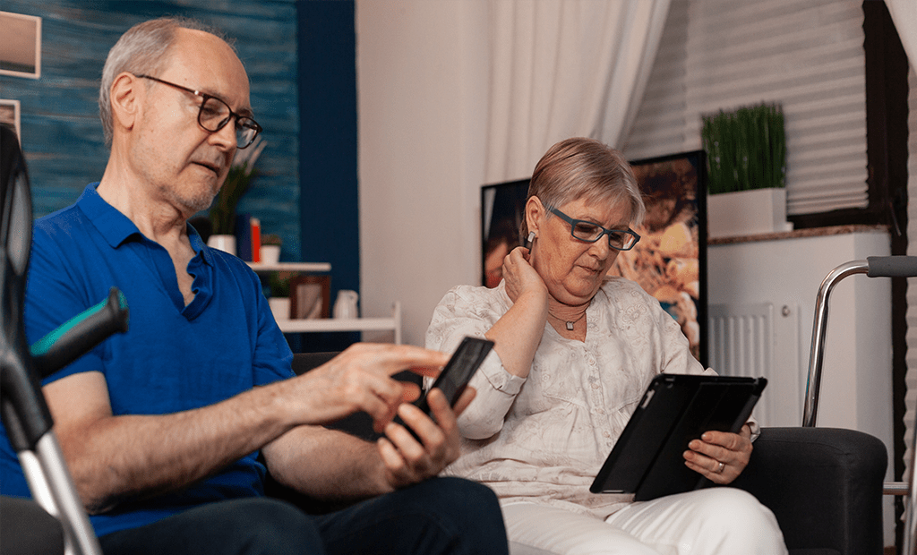 Acessibilidade. Imagem de um casal de idosos sentado na sala mexendo em aparelhos eletrônicos. O homem está mexendo em um celular e a mulher mexendo em um tablet.