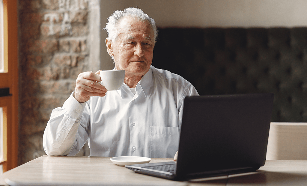 homem branco de cabelos grisalhos tomando café em frente a um computador