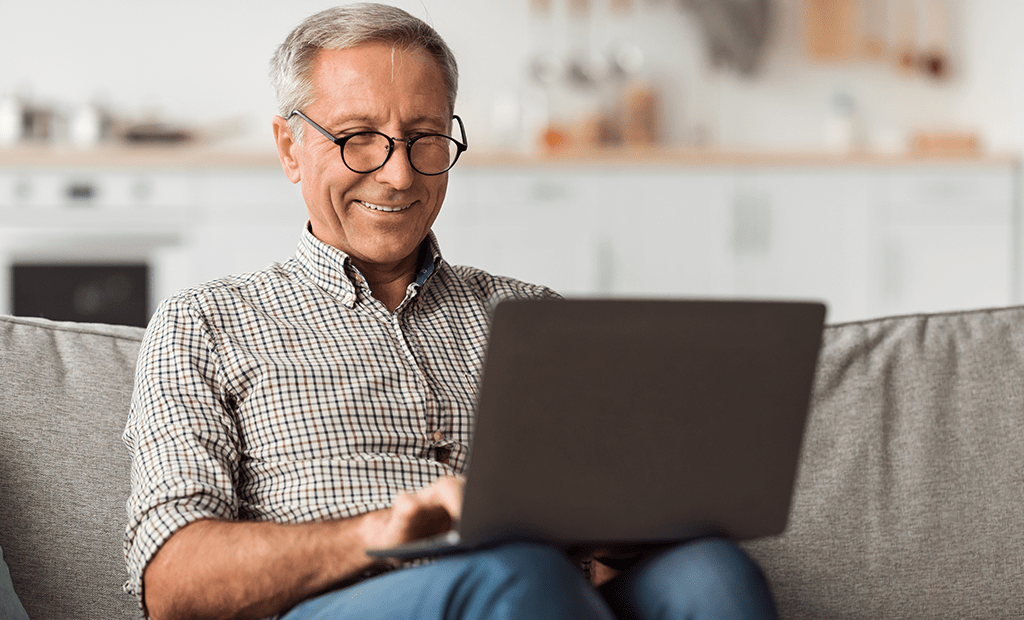 Acessível.  Imagem de um senhor de idade de cabelos grisalhos sorrindo e usando óculos, ele está sentado no sofá com um notebook em sua perna. 