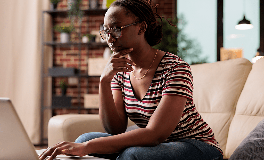 Libras. Imagem de uma mulher negra, sentado no sofá e mexendo no notebook. Ela usa óculos e cabelos preso.