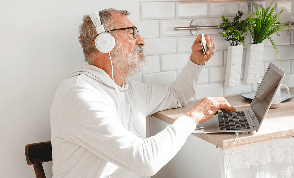 Acessibilidade. Imagem de um homem aparentando mais de 50 anos usando fones de ouvido. Em uma mão, ele digita no notebook e, na outra, segura um cartão.