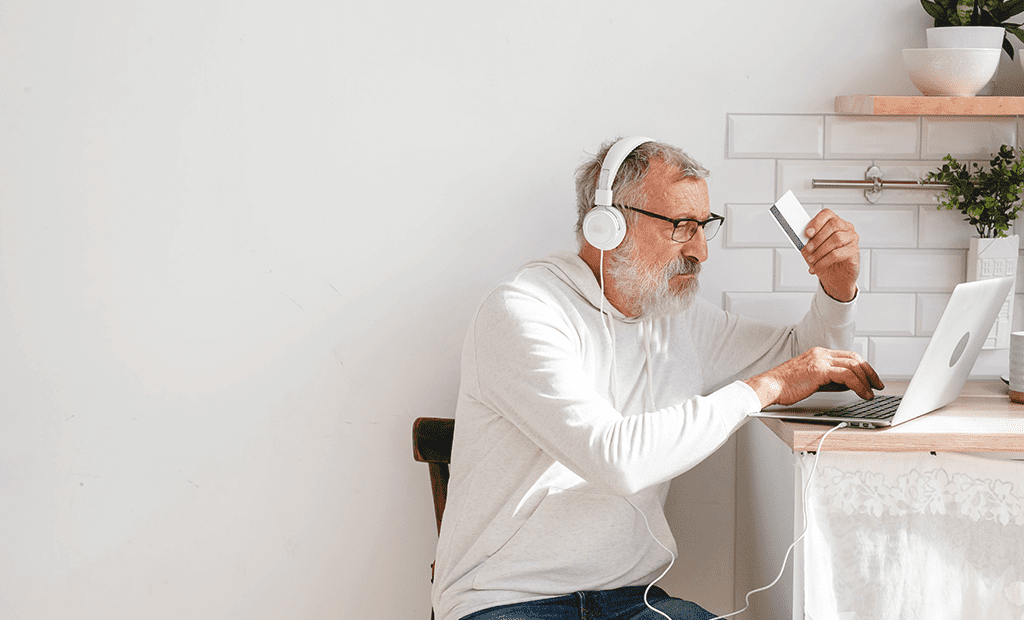  e-commerce. Imagem de um homem de cabelos grisalhos e usando óculos. Ele segura em uma das mãos um cartão enquanto mexe no notebook, 