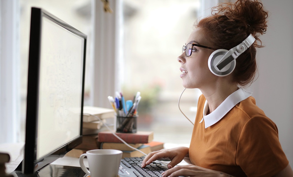 Siglas. mulher de cabelos ruivos presos em um coque, usando um par de fones de ouvido brancos e óculos. Ela está sentada em frente a um monitor de computador, digitando em um teclado.