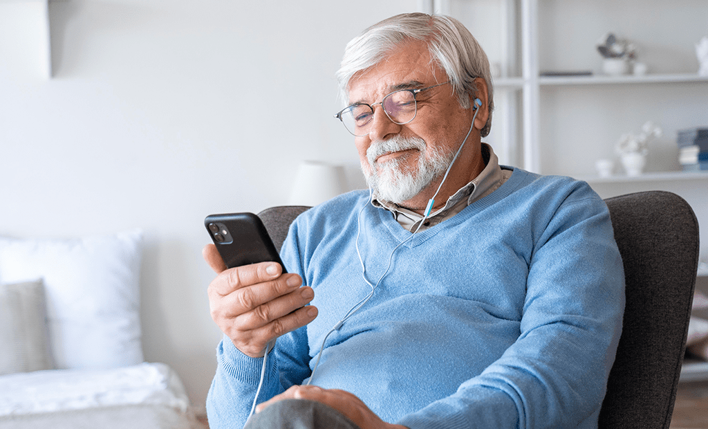 WCAG 3.0. Homem idoso sentado em uma cadeira, usando fones de ouvido conectados a um smartphone que ele segura. Ele veste uma blusa azul clara e óculos, aparentando estar em um ambiente doméstico. 