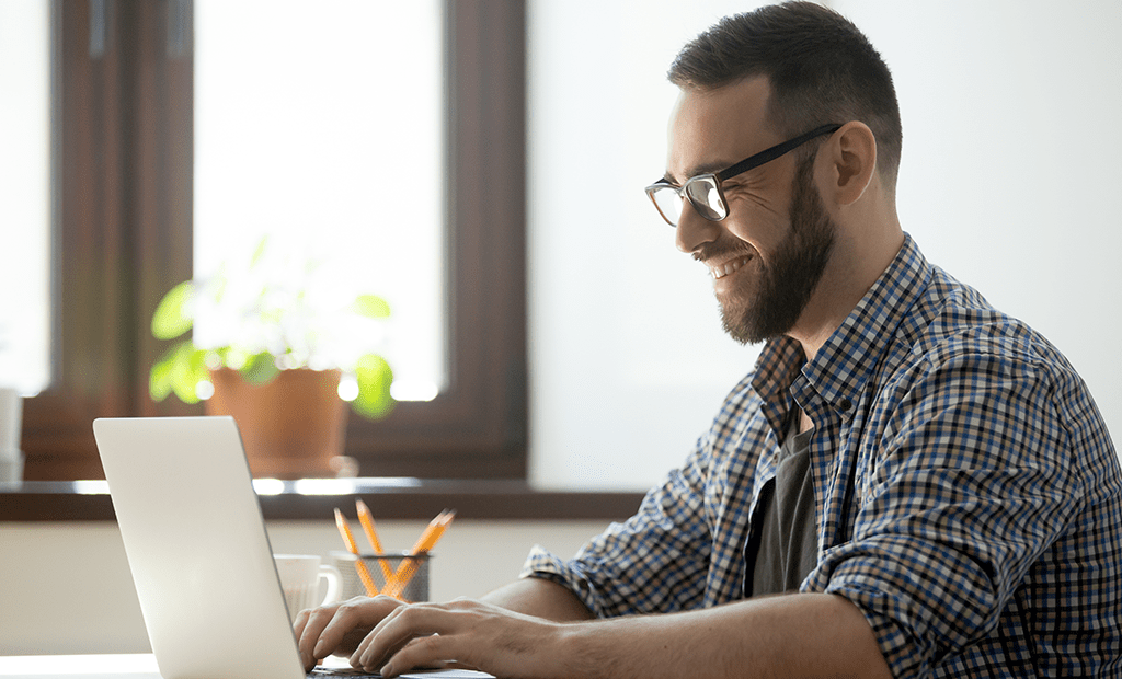 WCAG 3.0. Homem jovem usando óculos e digitando em um laptop. Ele está em um ambiente próximo a uma janela. O homem veste uma camisa xadrez.