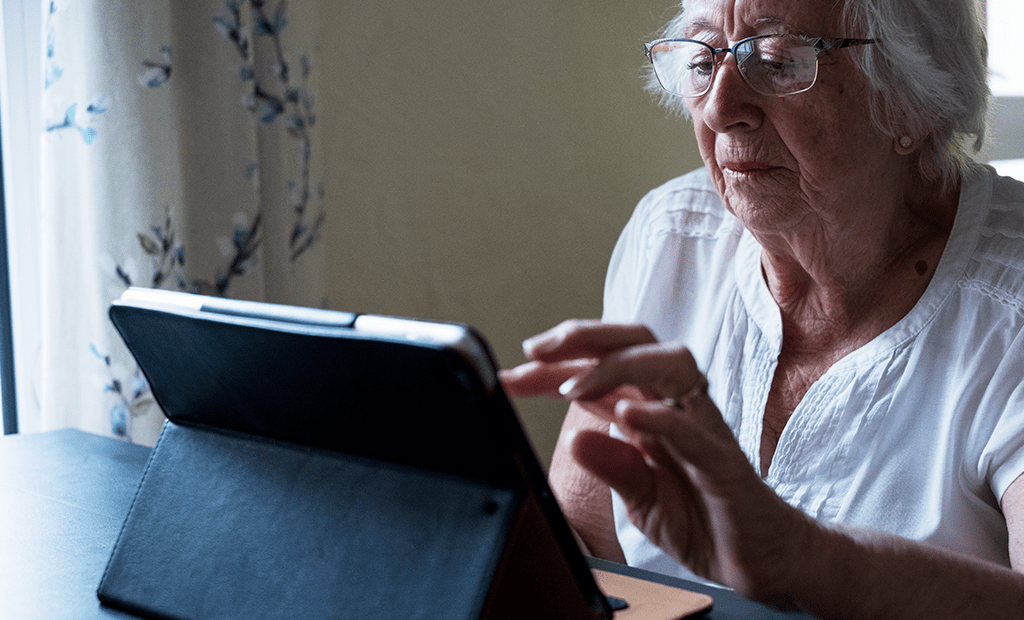 e-commerce. Mulher idosa, com cabelos brancos e óculos, usando um tablet. Ela está sentada à mesa, concentrada na tela do dispositivo, com as mãos próximas ao tablet.