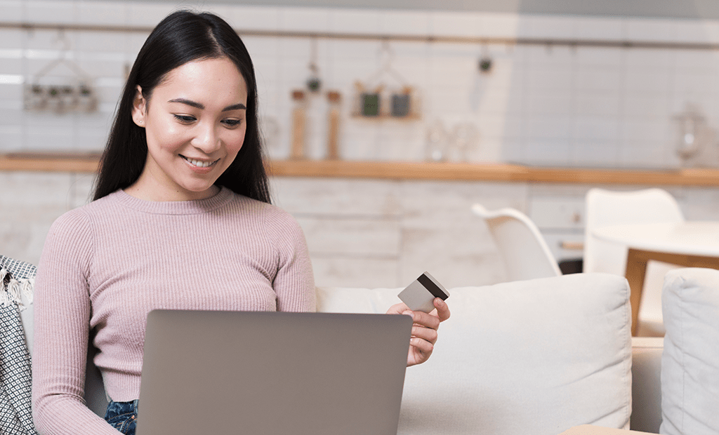  e-commerce. Uma jovem mulher sorridente, sentada em um sofá, segurando um cartão de crédito enquanto usa um laptop.