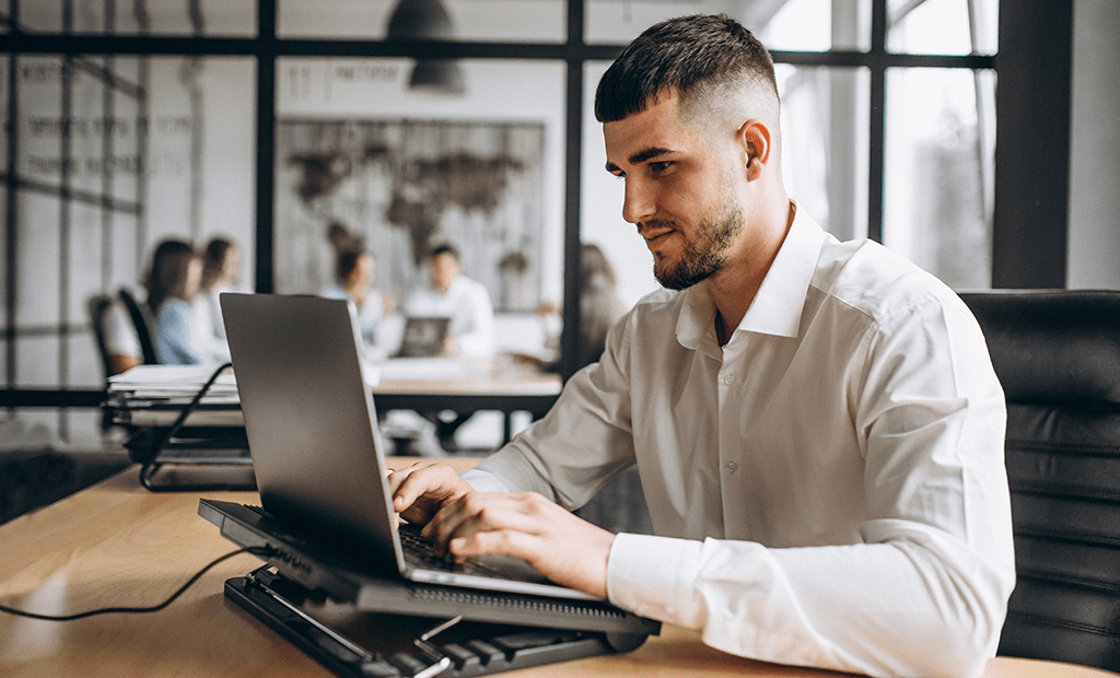 ESG. Homem jovem, com barba curta e cabelo escuro, vestindo uma camisa social branca, trabalhando em um laptop. Ele está sentado em uma mesa de escritório, concentrado na tela do computador. Ao fundo, desfocado, há um grupo de pessoas em uma reunião, sentado ao redor de uma mesa, conversando.