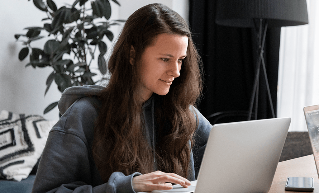 ESG. Mulher jovem com cabelos castanhos longos, concentrada em seu laptop. Ela veste um moletom cinza. 