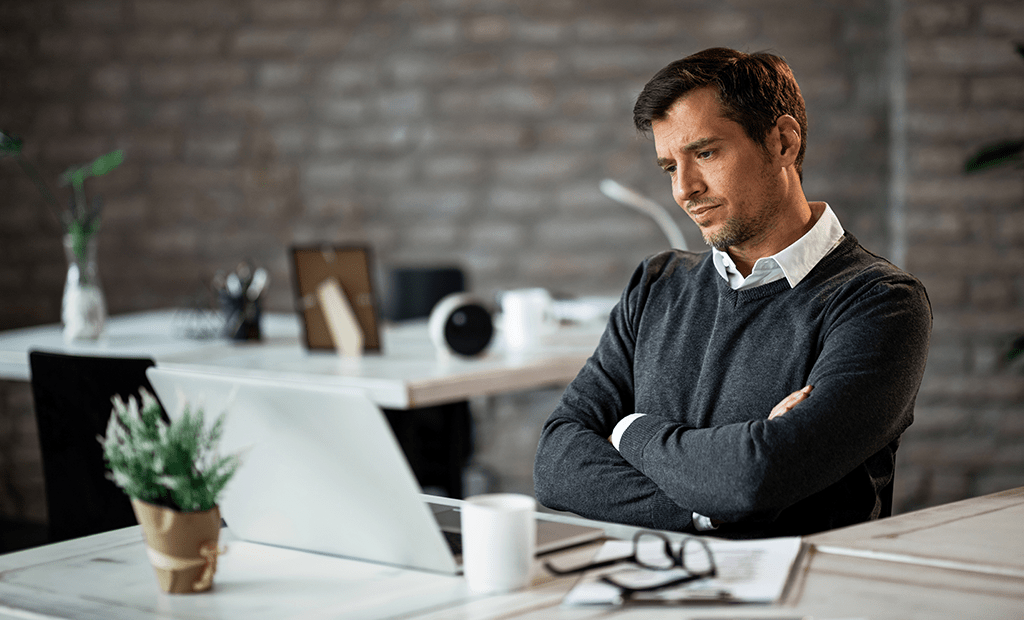 ESG. Homem adulto, de expressão pensativa, olhando para um laptop em uma mesa de escritório. Ele veste uma camisa branca por baixo de um suéter cinza escuro e está com os braços cruzados. Na mesa, há um par de óculos sobre alguns papéis, uma caneca e uma pequena planta decorativa.