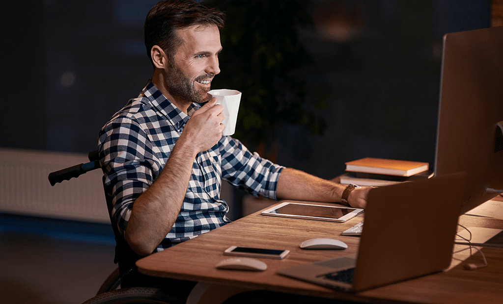 ESG. homem em uma cadeira de rodas trabalhando em um ambiente de escritório. Ele está vestindo uma camisa xadrez e parece concentrado enquanto olha para a tela de um computador, segurando uma xícara de café ou chá. Na mesa, ao lado dele, há um tablet, um smartphone e alguns livros, além de um teclado e um mouse.