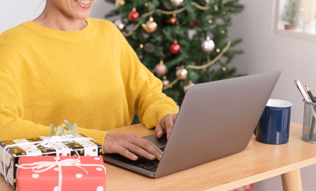 Black Friday. Pessoa sorrindo enquanto usa um laptop em uma mesa. Ela veste um suéter amarelo e está em um ambiente decorado para o Natal, com uma árvore de Natal ao fundo, adornada com bolas e enfeites. Sobre a mesa, há presentes embrulhados em papéis coloridos, uma caneca azul e um porta-lápis com alguns lápis.