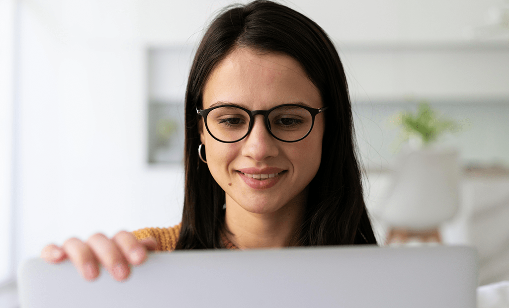 Black Friday. Mulher jovem, com cabelos castanhos e lisos, usando óculos de armação preta e brincos de argola dourados. Ela está sorrindo levemente enquanto olha para uma tela de computador.