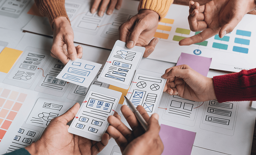 UX. Várias mãos de pessoas colaborando em um projeto de design de interface para um aplicativo ou site. Elas seguram diferentes cartões de protótipo, que contêm esboços de telas com elementos como caixas de texto, ícones e imagens.