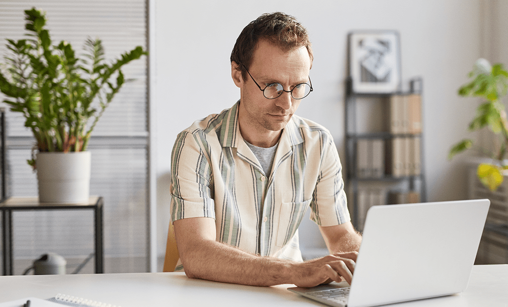 UX. Homem usando óculos, trabalhando concentrado em um laptop. Ele está em um ambiente bem iluminado e organizado, com plantas e estantes ao fundo.