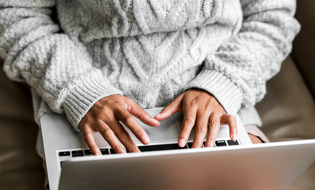 UX. Pessoa usando um laptop, com as mãos sobre o teclado. A pessoa está vestindo um suéter de lã cinza com detalhes trançados. 