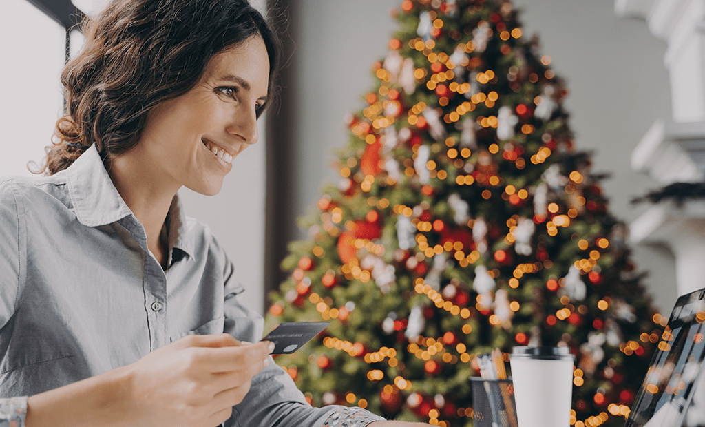 Fim de Ano. Em primeiro plano, uma mulher sorridente segura um cartão de crédito enquanto utiliza um laptop. Ela veste uma camisa cinza-clara e está em um espaço interno bem iluminado. Ao fundo, uma árvore de Natal decorada com luzes brilhantes, bolas e enfeites