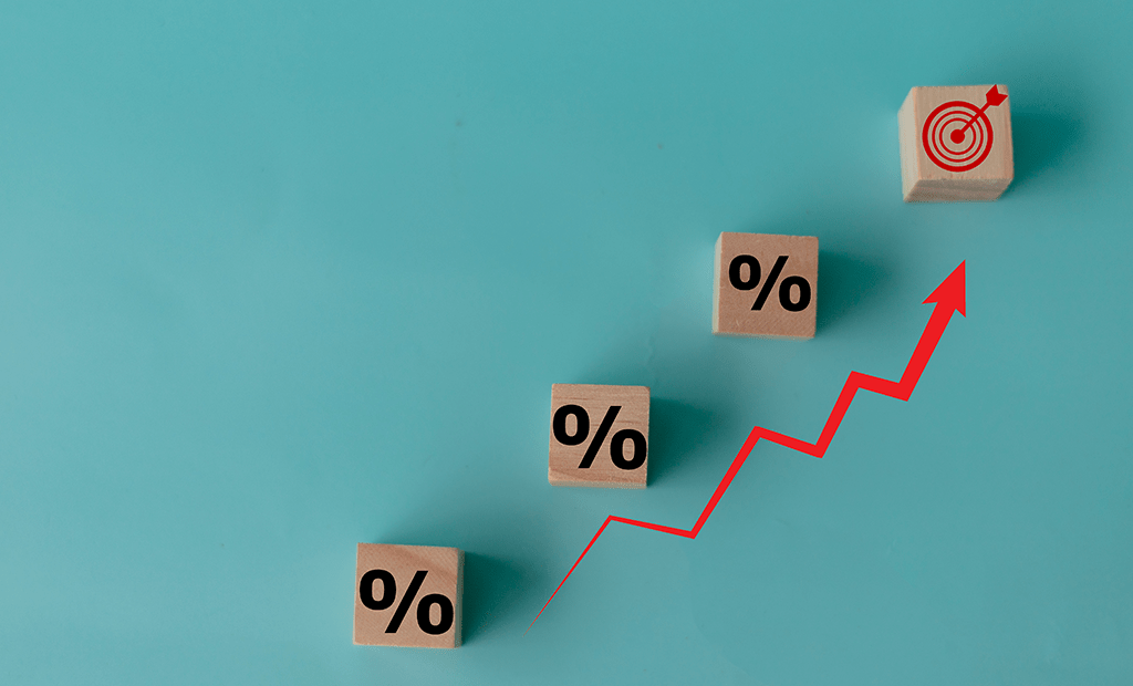 Fim de Ano. Quatro blocos de madeira estão dispostos em uma linha ascendente, simbolizando um progresso ou crescimento. Os três primeiros blocos têm o símbolo de porcentagem (%), enquanto o último exibe um alvo com uma flecha no centro, representando a conquista de uma meta. Uma linha vermelha em formato de gráfico de crescimento conecta os blocos.