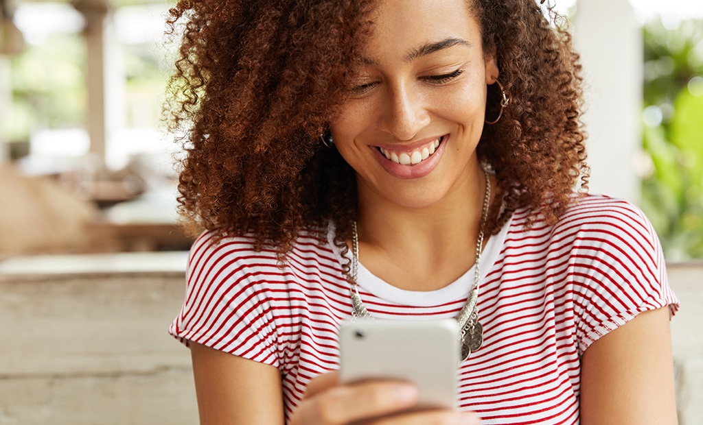 Calendário Inclusivo. mulher sorridente, com cabelos cacheados volumosos, vestindo uma camiseta listrada em vermelho e branco. Ela está segurando um celular branco e olhando para a tela com expressão de alegria. 