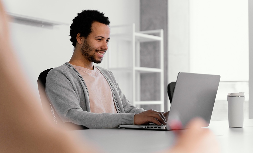 Calendário inclusivo. homem sentado em um ambiente de trabalho moderno. Ele está sorrindo e usando um laptop. O homem veste uma camiseta clara com um casaco cinza, e ao fundo há um ambiente claro e minimalista com prateleiras brancas. 