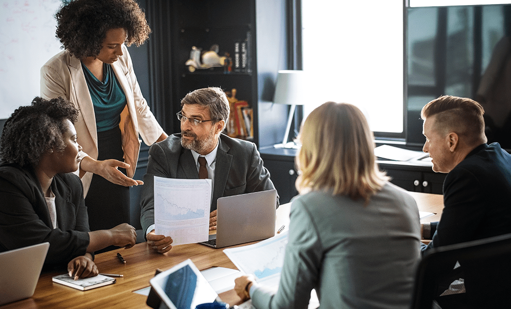 Acessibilidade Digital. Grupo de profissionais reunidos em uma sala de reuniões, discutindo documentos e gráficos. Um homem de terno e óculos segura um relatório enquanto conversa com os colegas. Uma mulher em pé gesticula.