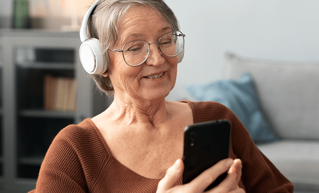 Acessibilidade Digital. Mulher idosa sorrindo enquanto usa fones de ouvido brancos e segura um smartphone. Ela parece estar assistindo a algo ou participando de uma videochamada. O ambiente ao fundo tem uma estante de livros e um sofá com almofadas.