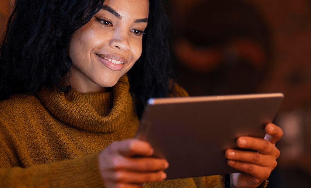 Acessibilidade Digital. Mulher sorridente segurando um tablet. Ela está vestindo um suéter de gola alta em tom mostarda e parece concentrada na tela do dispositivo.