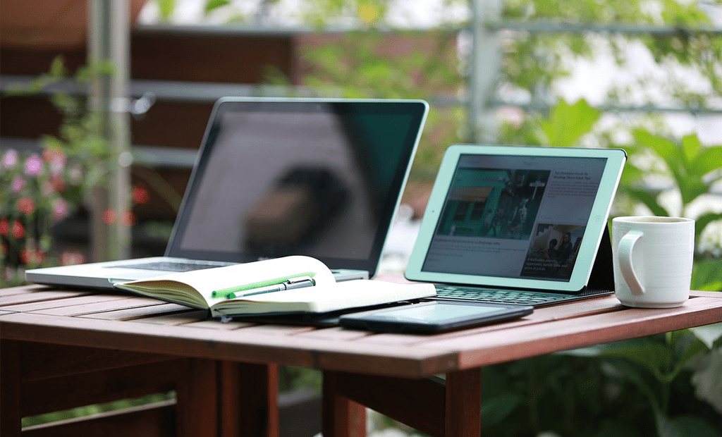 Termômetro. Mesa de madeira com um notebook, um tablet, um caderno aberto com caneta e uma caneca, em ambiente externo com plantas ao fundo.