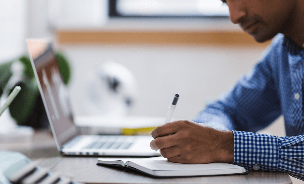 Termômetro. Pessoa escrevendo em um caderno ao lado de um notebook, em um ambiente de trabalho iluminado.