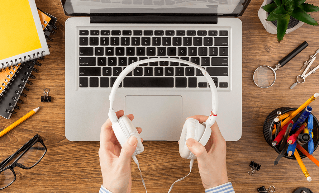 Leitores de tela. Mesa de trabalho com um notebook ao centro. Uma pessoa segura um fone de ouvido branco em frente ao teclado. Ao redor, há cadernos, lápis, canetas, uma lupa, óculos, uma planta e outros materiais de escritório.